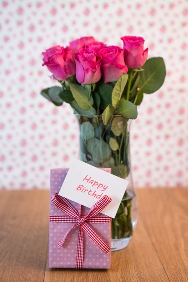 Bunch of pink roses in vase with pink gift leaning against it and happy birthday card