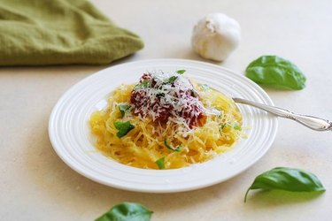 A serving of spaghetti squash topped with tomato sauce, basil and parmesan, in a white pasta bowl surrounded by bay leaves and a bulb of garlic