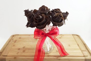 A vase on a wooden cutting board, tied with a red ribbon, containing a bouquet of dark Tootsie Roll roses.