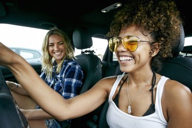 Portrait of two women with long blond and brown curly hair sitting in car, wearing sunglasses, smiling.