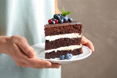 Woman holding plate with slice of chocolate sponge berry cake on grey background, closeup