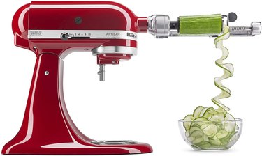 Red KitchenAid stand mixer with spiralizer attachment slicing a cucumber, against a white ground