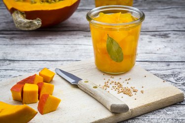 Glass of sweet-and-sour pickled pumpkin, mustard grains, knife and diced pumpkin on wooden board