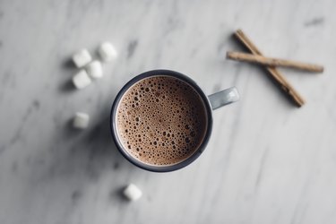 Overhead view of frothy hot chocolate in mug with marshmallows and cinnamon on table