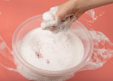 A Basin with Soapy Water, Hand Holds Soapy Sponge with Foam on Pink Background