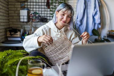 Young Blue haired woman knitting a wicker pot at home using laptop for watching online tutorial
