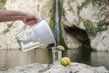 A hand pouring filtered water in the big glass with lemon on the nature background. Filter jug