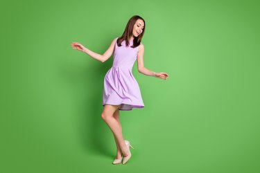 Full size photo adorable sweet careless girl lady look down hands side prom party dress air blow slim legs shoe shop advert concept wear purple dress isolated pastel green color background