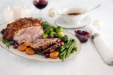 Roast pork ham with crust, green beans, Brussel sprouts, carrots, cranberry and sauce on white table cloth with candles and Christmas decoration, festive dinner, selected focus