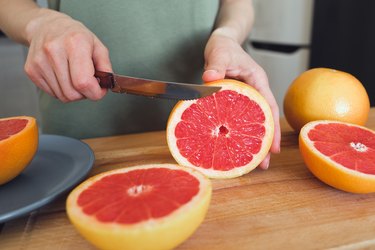 A girl or woman is holding a knife, cutting a red ripe grapefruit in half, on a cutting board, against the background of a wooden kitchen table. There is a manual juicer nearby. Making juice for breakfast. The concept of vegetarian, vegan and raw food.