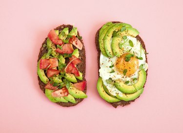 Toasts of dark bread with avocado slices, red tomatoes, fried egg and microgreens, against pink background