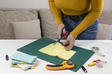 woman in a yellow sweater cuts the fabric. there are quilting tools on the table. patchwork knife, scissors, lined cutting mat, self-locking, thread, measuring tape. sewing supplies