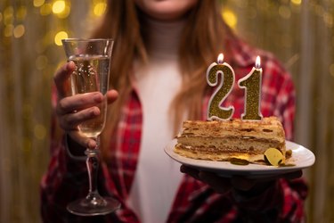 Hands of young woman holding birthday cake with number 21 candles and glass of champagne celebrating birthday.