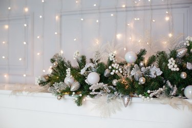 fireplace is decorated with white and silver balls, bells, cones and Christmas tree wreath. Christmas garland on mantelpiece. Christmas wreath and decorations on fireplace with sparkling garland.