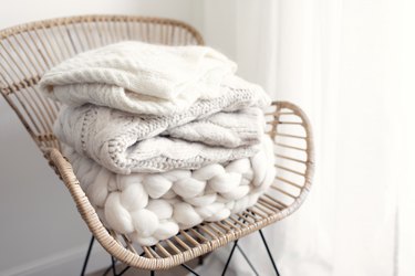 Stack of cozy wool white knitted sweaters on wicker armchair. Pile of merino and cashmere winter clothes. Hygge style. Warm concept.