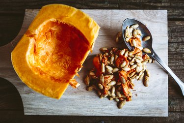 Hollowed-out pumpkin and its seeds on a wooden cutting board, on top of a rustic wood surface