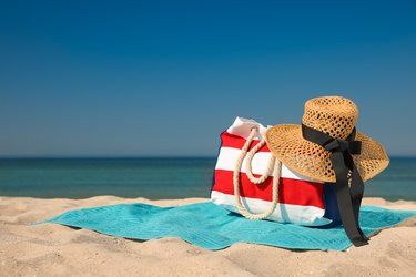 Turquoise beach towel with sun hat and white and red striped beach bag on sand near sea.