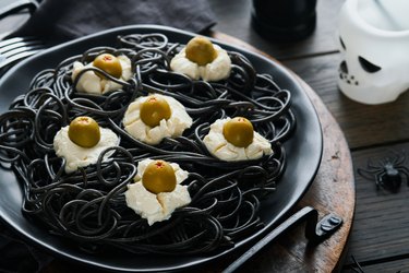 Halloween party Italian black pasta decorated horror  olives like eyes on black plate on old dark table background. Monster face from pasta. Halloween decorations and Halloween food concept. Mock up.