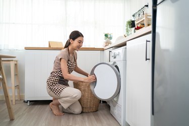 Asian beautiful woman put dirty clothes to washing machine in house. Attractive girl wear apron sit on floor, feel happy to loads laundry in washer appliance at home. Domestic-House keeping concept.