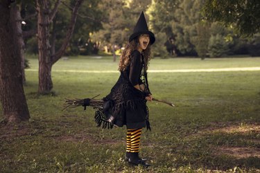 Little girl in witch costume and black long hat holding wooden broom, standing outdoor against autumn forest background.