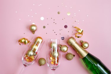 Green and golden glass bottles of champagne, two glasses and golden confetti over pink background. Festive New Year Greeting Card.