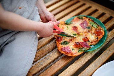 Unrecognizable small child cutting homemade pizza at home with scissors.