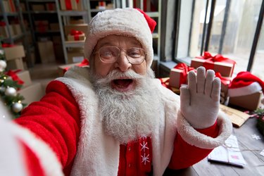 Happy old bearded Santa Claus wearing costume holding phone waving hand taking selfie, video calling, recording video Merry Christmas greeting or shooting vlog standing in workshop, face camera view.