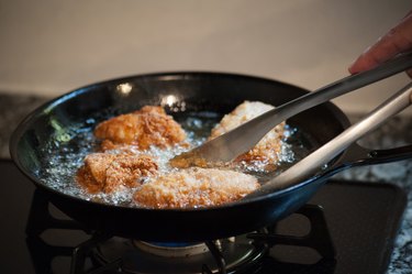 Fried chicken cooking in a cast-iron skillet on a gas stove, with the cook's hand using tongs to touch a piece of chicken
