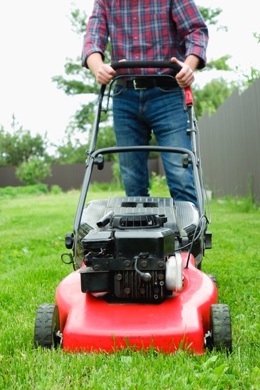 Lawn grass mowing. A man in a plaid shirt and blue jeans mows the grass with a lawn mower.