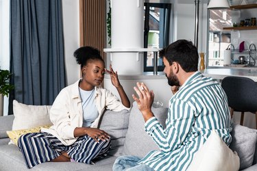 Young stressed and angry woman sitting on the sofa with displease facial expression arguing with her boyfriend about his bad habit of smoking cigarettes. Stop smoking concept.