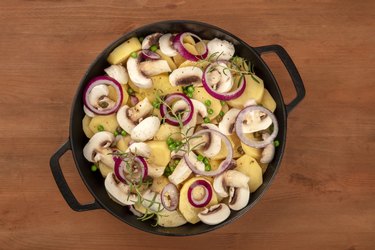 A top-down photo of a braiser with raw vegetables, sliced potatoes, onions, green peas, mushrooms, and rosemary, shot from above on a dark rustic wooden background wih copy space