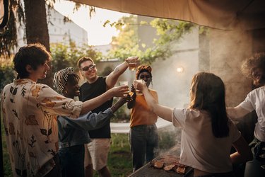 Friends raise their glasses to cheers as they stand around an outdoor grill on a sunny day.