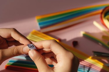 Paper and quilling tools on a pink background with hands and space for text and design.