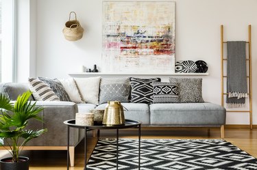 Patterned pillows on grey corner sofa in living room interior with table and painting. Real photo