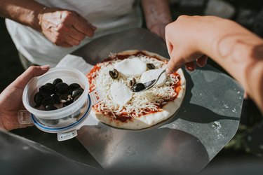 Hands adding black olives to the top of a home-made pizza