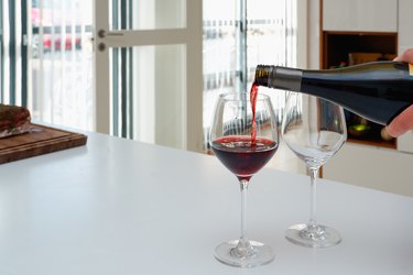 A woman pours red wine from a bottle into wine glasses. Modern white kitchen clean interior design
