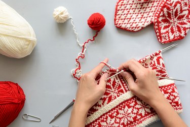 Girl knits red-and-white Norwegian jacquard hat with knitting needles against gray background
