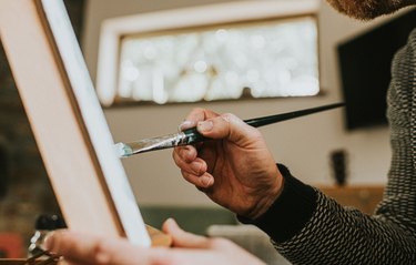 A man wearing glasses concentrates as he paints with oils onto a canvas on an easel