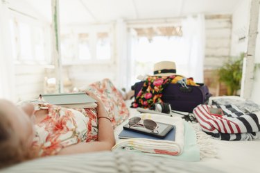 Woman with book relaxing on bed next to suitcase in sunny beach hut bedroom