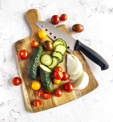 Knife with sliced salad ingredients (tomatoes, cucumber and onion) on cutting board on white marble background