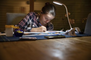 Teenager doing paint by numbers art at a dining table in low light in the evening, under a portable desk lamp and wearing wireless in-ear headphones.