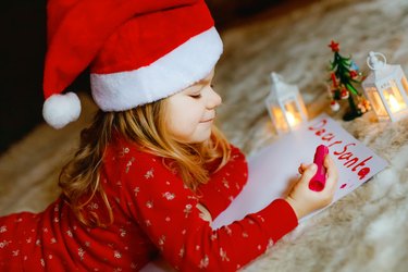 Little cute preschool girl in pajamas under Christmas tree, writing wish list letter to Santa Claus at home, indoors. Traditional Christian festival. Happy toddler child waiting for gifts on xmas.