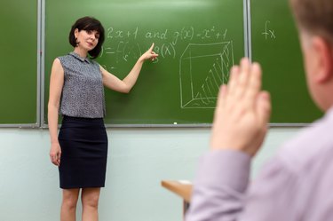 The teacher at the blackboard with a smile asks the answer from the student who pulls his hand