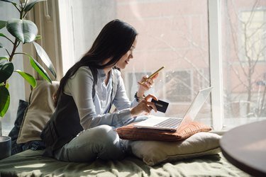 A middle-aged asian woman in blue jeans sitting on the bed in a yoga pose in front of a laptop