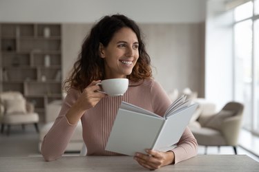Happy young hispanic woman reading book and drinking coffee.
