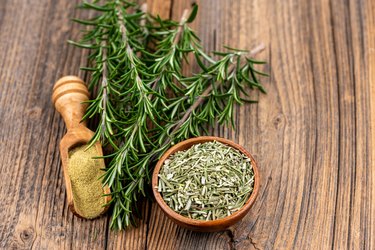 A wooden bowl filled with whole dried rosmary, a spice shovel with ground rosemary and two rosemary twigs on a rustic wooden background