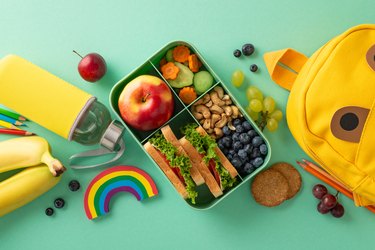 lunchbox filled with fresh food, cookies, water, stationery, colored pencils, rainbow plasticine, funny backpack on teal background