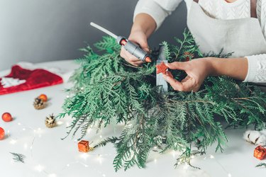 Florist woman makes Christmas composition for table decoration from thuja, spruce branches in wooden box. Glue hot gun, star, cones, snow apples, balls, cotton flowers,garland. New Year celebration