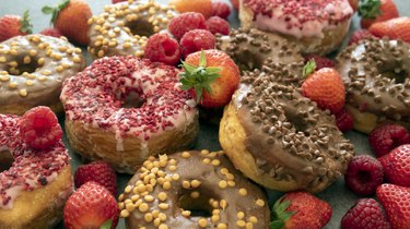 Full frame image of heap of glazed ringed Cronuts (croissant-doughnut pastry) with chocolate or strawberry fondant icing and sprinkles, fresh strawberries and raspberries, blue background, elevated view, focus on foreground