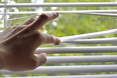 man's hand opens strips of blinds to look through the window